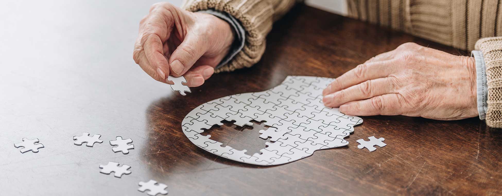 Hände einer älteren Person, die einen Menschenkopf puzzelt; der mittlere Teil des Kopfes ist ohne Puzzleteile