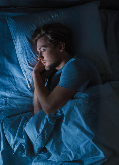 Draufsicht auf einen jungen Mann, der nachts gemütlich auf einem Bett in seinem Schlafzimmer schläft. Blaue nächtliche Farben mit kaltem, schwachem Laternenlicht, das durch das Fenster scheint.