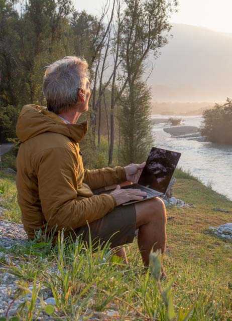 Mann, der am Laptop arbeitet, entspannt sich bei Sonnenaufgang in der Nähe des Flusses