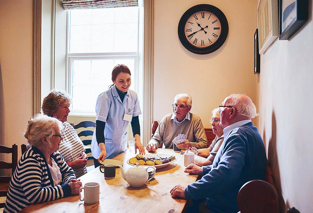 Ältere Personen sitzen an einem Tisch, eine Pflegerin bringt Kaffee und Kuchen