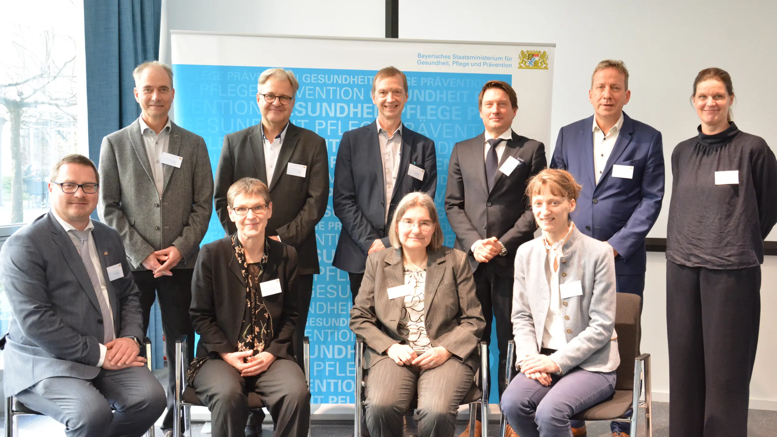 Gruppenfoto mit 11 Teilnehmern (4 sitzend, 7 stehend) vor Banner des Bayerischen Staatsministeriums für Gesundheit, Pflege und Prävention